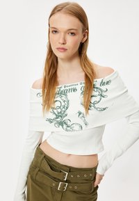 Weiße, schulterfreie, gerippte Bluse mit grünem Grafiktext und floralen Designs, kombiniert mit olivgrünen, hochgeschnittenen Hosen mit Gürtel.