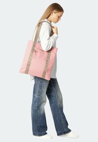 Pink fabric tote bag with a rectangular shape, featuring beige straps with text and a front zip pocket. Worn by a person in casual attire.
