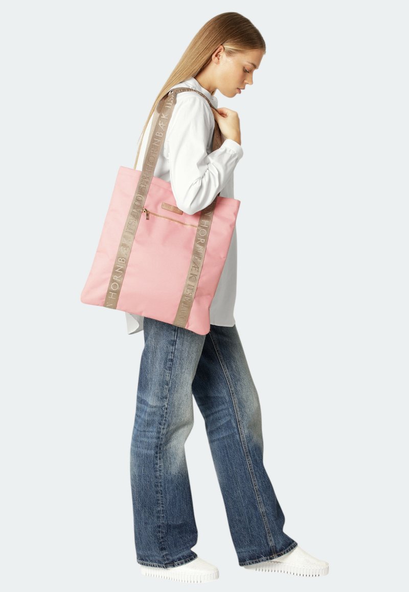 Pink fabric tote bag with a rectangular shape, featuring beige straps with text and a front zip pocket. Worn by a person in casual attire.