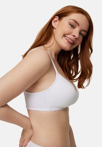 Sujetador blanco con tirantes finos, diseño sin costuras, tejido suave y detalle de borde con borde ondulado. La modelo posa de lado, mostrando el ajuste y la forma.