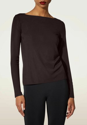 Donna che indossa una maglia aderente a maniche lunghe marrone scuro con una ampia scollatura rotonda e pantaloni neri su uno sfondo chiaro.