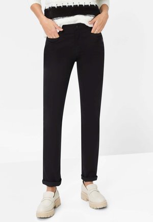 Pantaloni neri su misura con taglio dritto, dotati di tasche frontali e risvolti arrotolati. Abbinati a mocassini chunky di colore chiaro.