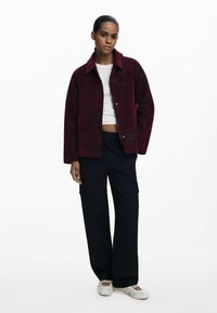 Giacca in velluto a coste bordeaux con colletto, bottoni frontali e tasche laterali, abbinata a pantaloni cargo neri larghi e scarpe bianche slip-on.