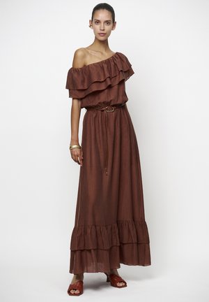 Vrouw draagt een mouwloze, off-shoulder bruine maxi-jurk met ruches en een dunne riem, gecombineerd met bruine sandalen met hak en een gouden armband.