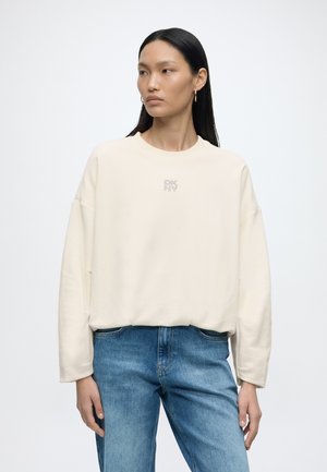 Kvinde med langt mørkt hår iført guldringeøreringe, en offwhite sweatshirt med lille logo og blå jeans, stående mod en ensfarvet baggrund.