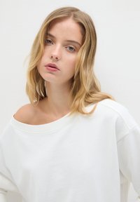 Giovane donna con capelli biondi che indossa un felpa bianca con spalle scoperte, che guarda la macchina fotografica con un'espressione neutra su uno sfondo semplice.