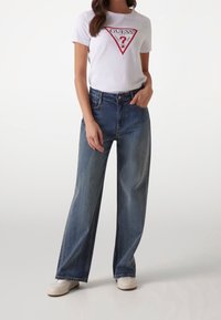 Camiseta blanca con logo rojo, combinada con jeans de pierna ancha de color azul claro. Los jeans presentan una cintura alta y costuras estándar, complementados con zapatillas blancas.