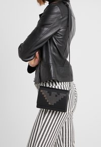 Žena v černé kožené bundě a pruhovaných kalhotách černobílé barvy nese černou kabelku přes rameno s kovovými hroty.