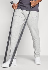 Pantaloni de trening gri din bumbac cu manșete elastice, având un logo brodat. Purtați cu un tricou gri ajustat și adidași albi, ținând benzi de rezistență.
