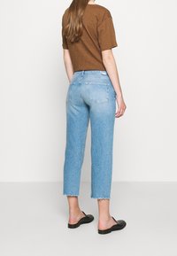 Vaqueros azules claros recortados con bajos deshilachados combinados con una camiseta marrón y zapatos negros tipo slip-on, vistos desde atrás.