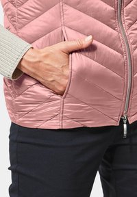 Gevoerd vest in rozenpink, met een quilted ontwerp, een rits aan de voorkant en een zijzak. Daaronder zijn donkerblauwe broeken gedeeltelijk zichtbaar.