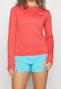 Nainen, joka on pukeutunut korallinväriseen Nike-pitkähihapaiseen paitaan ja vaaleansinisiin urheilushortseihin, seisoo rentoina sivuilla käsivarsin yksinkertaisen taustan edessä.