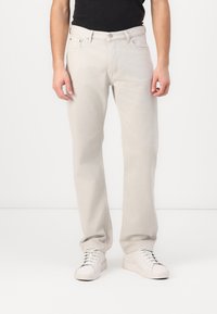 Jeans de mezclilla beige con pierna recta, cinco bolsillos y presillas para cinturón, combinados con zapatillas blancas sobre un fondo neutro.