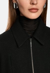 Schwarzer Wollmantel mit kragenförmigem Ausschnitt und Reißverschluss, kombiniert mit einem transparenten schwarzen Rollkragenpullover und silbernen Statement-Ohrringen.