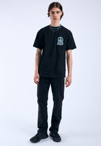 Camiseta de algodón negra con un diseño gráfico azul claro en el pecho, combinada con pantalones vaqueros oscuros y zapatillas negras gruesas.