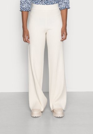 Femme portant un pantalon crème à taille haute et jambes larges côtelé, avec des chaussures épaisses crème, debout devant un fond clair uni.