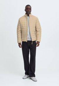 En person står selvsikkert iført en tan quiltet jakke over en grå sweater, parret med mørke bukser og sneakers mod en lys baggrund.