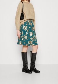 Beige ribbed sweater, hame tealiana bunga bercetak emas, dan sepatu bot kulit hitam setinggi lutut, dilengkapi dengan tas kecil hitam.