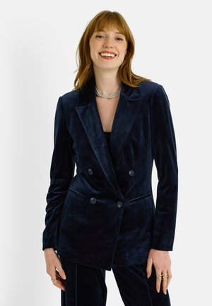 Blazer cruzado de terciopelo azul marino con solapas en punta, dos bolsillos frontales y botones negros. Textura suave y ajuste entallado.