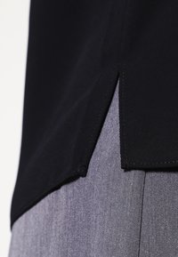 Svart tygblus med rak nederkant, med en kontrasterande grå texturerad nederdel. Inga synliga mönster eller detaljer i hårdvara.