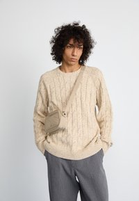 Joven con cabello rizado, vistiendo un suéter beige de punto trenzado, pantalones grises de rayas y un pequeño bolso cruzado beige, con las manos en los bolsillos.