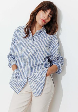 Femme aux cheveux bruns portant une chemise à manches longues bleu et blanc rayée avec des motifs blancs, et un pantalon de couleur claire, posant contre un mur blanc.