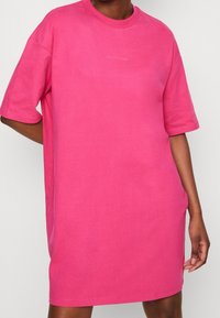 Vaaleanpunainen ylisuurennettu t-paita-mekko, joka on valmistettu pehmeästä kankaasta. Mekossa on pyöreä pääntie, lyhyet hihat ja rintakehässä logo.