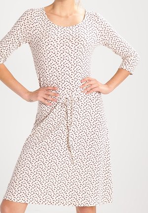 Woman wearing a white knee-length dress with small red floral pattern, three-quarter sleeves, and a tied waist belt, hands resting on hips.