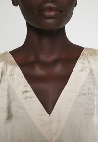 Top di seta beige con un design a V profonda, che mostra una consistenza liscia e una leggera lucentezza, adagiate dolcemente su una pelle scura.