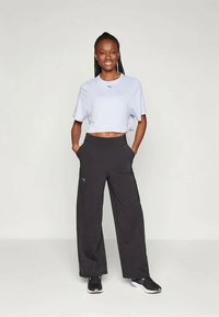 Smaidoša sieviete stāv ar rokām kabatās, uzvilkusi baltu īsu T-kreklu, brīvus melnus bikses un melnas sporta apavus uz vienkrāsaina fona.