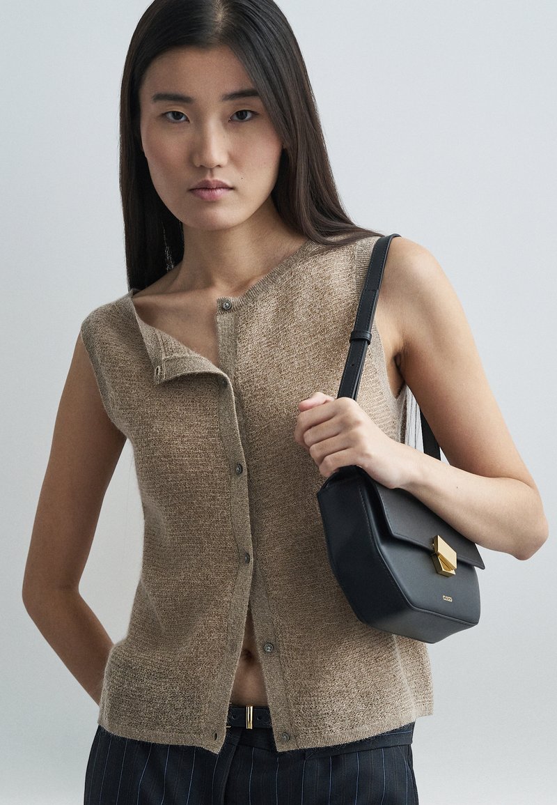 Beige gestricktes ärmelloses Oberteil mit einem Knopfverschluss und asymmetrischem Ausschnitt, kombiniert mit einer schwarzen Lederhandtasche mit einem goldenen Verschluss.