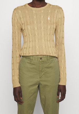 Persona que lleva un suéter beige de punto trenzado con logo en el pecho y pantalones verde oliva de cintura alta, de pie contra un fondo liso.