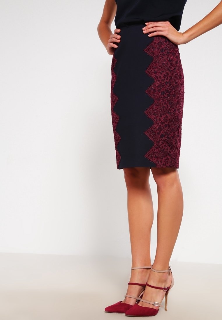 Femme portant une jupe crayon noire avec des panneaux en dentelle bordeaux en zigzag, associée à des chaussures à talons hauts bordeaux et beige avec bride à la cheville.