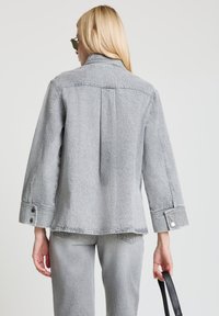 Graue Denimjacke mit Kragen, einer Rückenfalte und Knöpfen an den Ärmelabschlüssen. Sie hat eine lockere Passform und wird mit passenden grauen Hosen kombiniert.