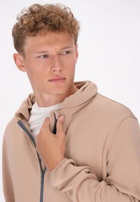 Beige rits-hoodie voor heren, gemaakt van gestructureerde stof, met een kangoeroezak en grijze rits. Voorzien van een verstelbare trekkoordkap.
