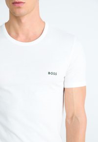T-shirt en coton blanc avec un col rond. Présente un petit logo "BOSS" brodé en vert sur la poitrine gauche. Manches courtes.