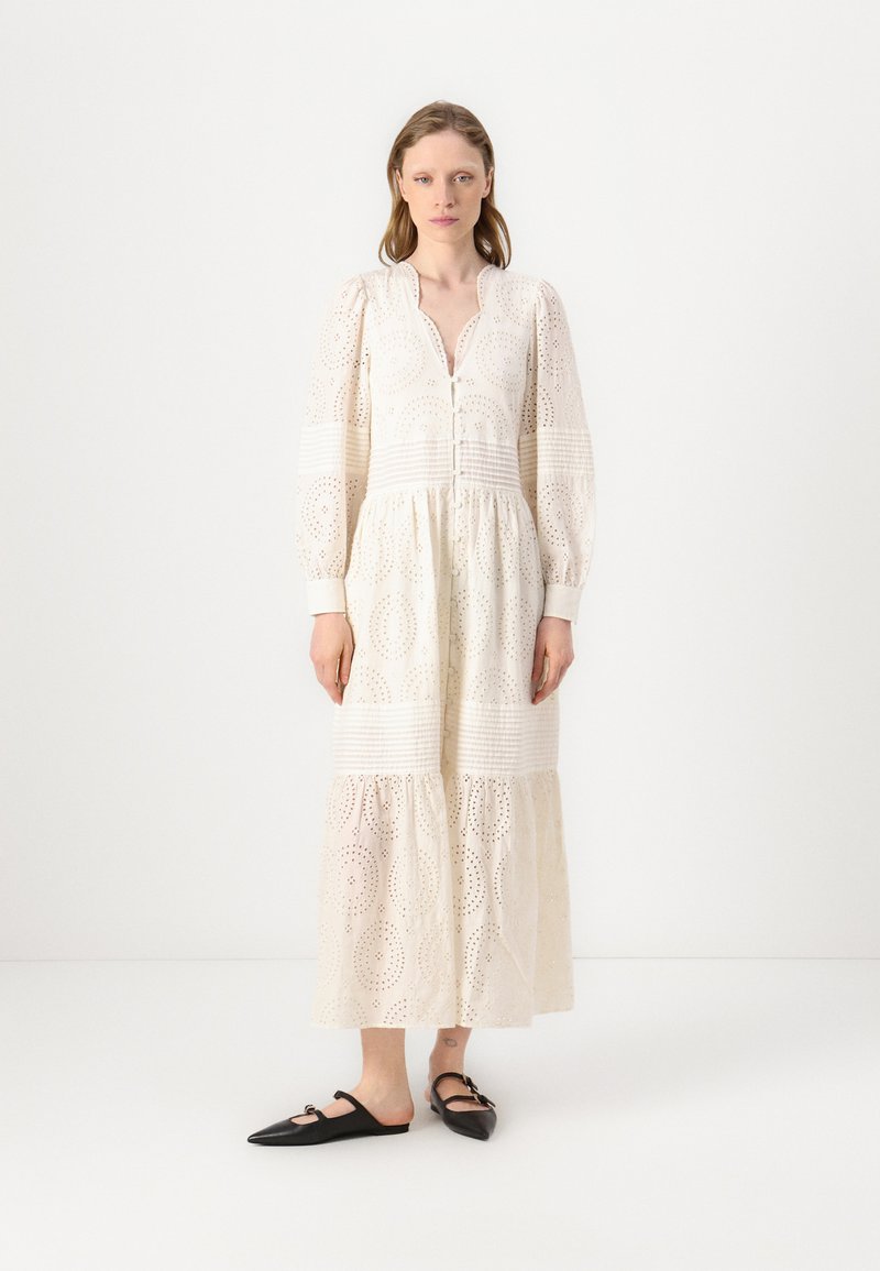 Hvit langermet kjole med broderi anglais-detaljer, knappfront og utsvingt skjørt. Kombinert med svarte slip-on sko.