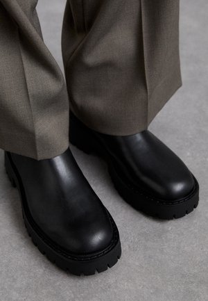 Bottes épaisses en cuir noir portées avec un pantalon vert olive sur une surface grise, montrant la texture des chaussures et les détails de l'ourlet du pantalon.