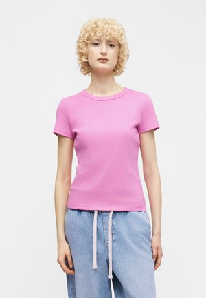 Personne aux cheveux blonds bouclés et courts, portant un t-shirt rose ajusté à manches courtes et un jean bleu clair avec des cordons de serrage roses, sur un fond blanc.