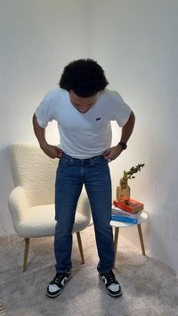 White short-sleeve shirt, blue denim jeans, black and white sneakers. Indoor setting with a textured background, a chair, and decorative items.