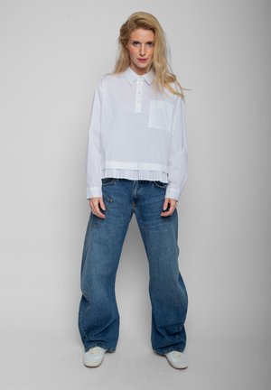 Blonde Frau steht vor grauem Hintergrund, trägt ein weißes Langarmshirt, lockere blaue Jeans und weiße Sneakers.