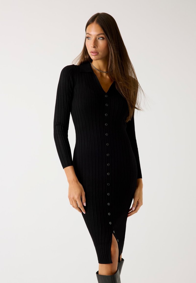 Abito midi nero a coste con scollatura a V, maniche lunghe e chiusura frontale con bottoni. Presenta una spacco alto e una texture liscia.