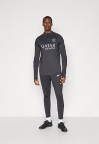 Αθλητική εμφάνιση σε μαύρο χρώμα, περιλαμβάνει μπλούζα με μακριά μανίκια, φερμουάρ και λογότυπο, συνδυασμένη με εφαρμοστά παντελόνια και μαύρα αθλητικά παπούτσια.