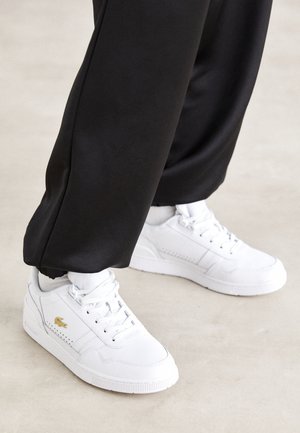 Weiße Low-Top-Sneaker mit goldenem Logo, getragen mit schwarzen Hosen und weißen Socken auf einem hellen, glatten Boden.