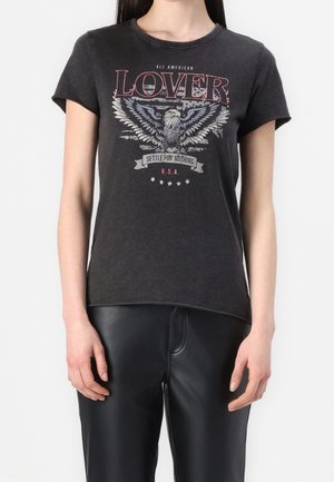 Frau trägt ein schwarzes Grafik-T-Shirt mit Adler und dem Text „ALL AMERICAN LOVER, SETTLE FOR NOTHING, U.S.A.“ und schwarze Lederhose.