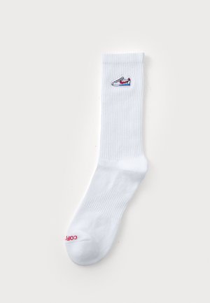 Baltā kokvilnas zeķe ar ribota raksta virsmu, kurai ir mazs izšūts kedu logs un sarkans teksts pirksta daļā.