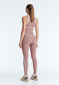 Femme avec queue de cheval brune portant une tenue de sport mauve sans coutures avec des accents blancs et des baskets beiges, debout devant un fond blanc, vue de dos.