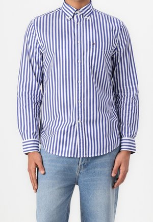 Homme portant une chemise boutonnée à rayures verticales bleues et blanches avec un petit logo sur la poche poitrine et un jean bleu clair.