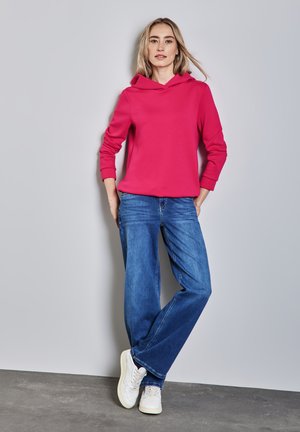 Sweat-shirt à capuche rose vif, associé à un jean en denim bleu à jambes évasées et des baskets blanches. Tissu lisse et coupe décontractée.
