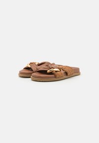 Brown slide sandals made of soft material, featuring woven accents and a large gold-tone hardware detail. Flat sole with textured border.